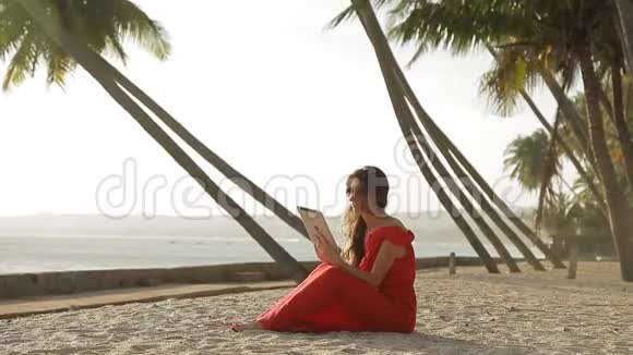 穿着红色裙子走在海边的年轻女子她在用平板电脑微笑着视频的预览图