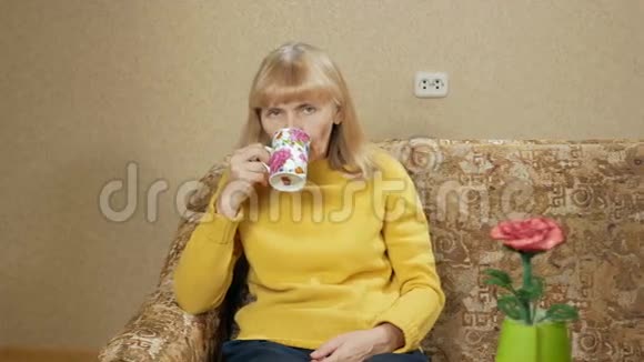 一个年老的女人在家里的沙发上喝着一杯热饮料看着相机3她辛苦了一番视频的预览图
