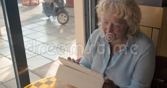 成熟的女人坐在咖啡馆里享受她的平板电脑在窗外的夏天阳光明媚的一天视频的预览图