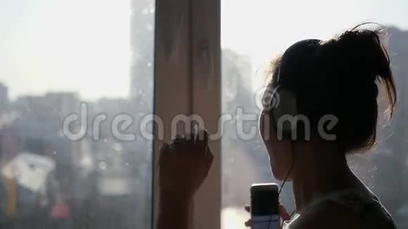 女孩欢快地戴着大耳机在城市背景的窗户附近跳舞手里拿着一部手机的阳光慢动作视频的预览图