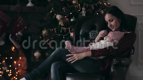 母亲坐在椅子上抱着一个正在睡觉的婴儿靠近圣诞树视频的预览图