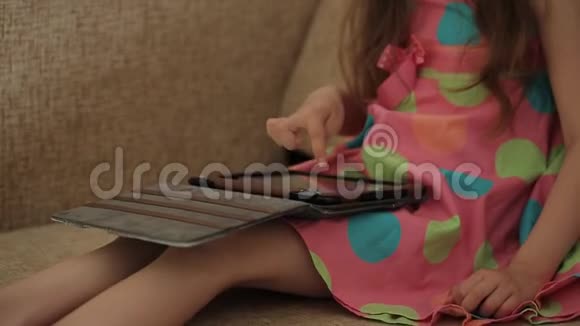 小女孩穿着一件漂亮的彩色连衣裙坐在靠窗的椅子上用平板电脑视频的预览图