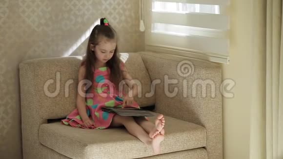 小可爱坐在沙发上玩平板电脑小女孩穿着漂亮的彩色裙子坐在椅子上视频的预览图