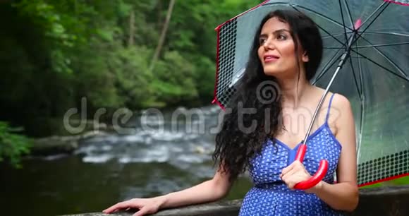 年轻的女人站在森林里带着一把伞靠近瀑布看着落水女人微笑着视频的预览图