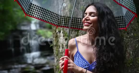 年轻的女人站在森林里带着一把伞靠近瀑布看着落水女人微笑着视频的预览图