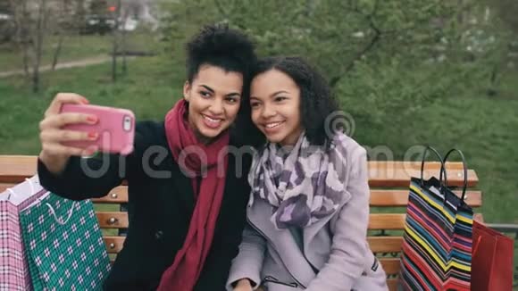 两个可爱的非裔美国女人带着购物袋和微笑在智能手机上自拍女朋友坐着视频的预览图