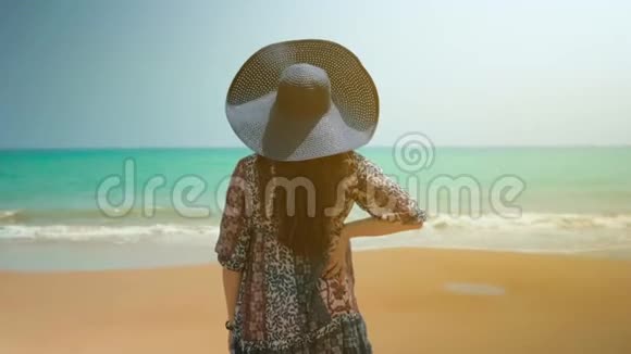 海滩上穿着蓝色连衣裙的女孩看着海洋摘下蓝色帽子近距离射击视频的预览图