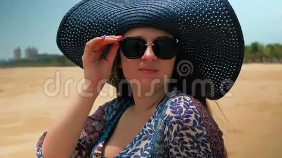 美丽的快乐女孩戴着蓝色的帽子和眼镜坐在阳光明媚的夏日海滩上近距离射击慢动作视频的预览图
