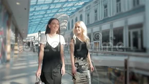 美丽的年轻时尚女性带着女朋友带着购物袋靠近商店橱窗在购物中心散步两个女孩视频的预览图