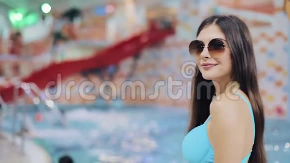 穿着蓝色泳衣和太阳镜在游泳池里摆姿势的年轻女性水上公园里一个漂亮女孩的肖像视频的预览图