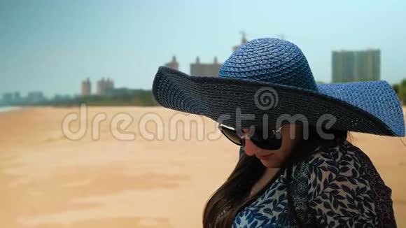 美丽的快乐女孩戴着蓝色的帽子和眼镜坐在阳光明媚的夏日海滩上近距离射击慢动作视频的预览图
