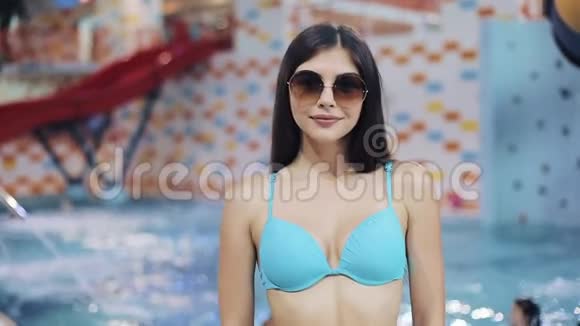 穿着蓝色泳衣和太阳镜在游泳池里摆姿势的年轻女性水上公园里一个漂亮女孩的肖像视频的预览图