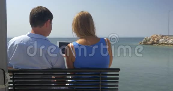 年轻人和年轻人坐在沙滩上的长椅上在海上天际线的背景下看着平板电脑和平板电脑上的东西视频的预览图