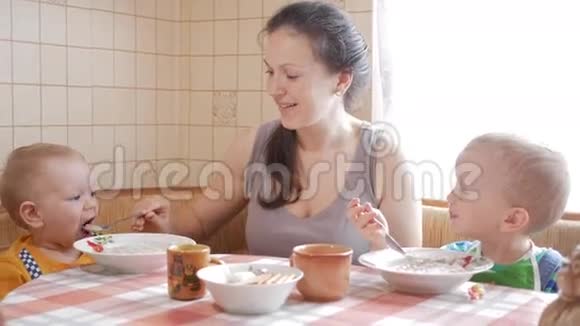 妈妈在桌旁给两个儿子喂粥一个男孩自己用勺子吃饭第二个孩子帮助一个女人视频的预览图