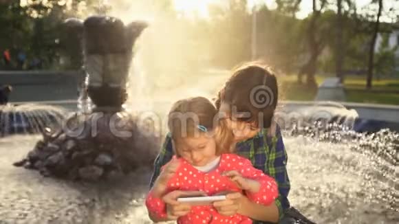 母亲和女儿在日落时在喷泉处用电话视频的预览图