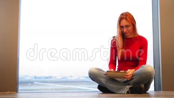 年轻漂亮的红发和戴眼镜的女人在机场的窗户附近使用小工具视频的预览图