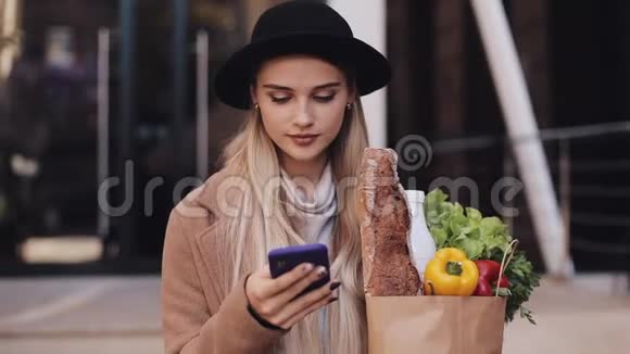 年轻漂亮的女人穿着时髦的外套走在街上拿着一包产品用智能手机视频的预览图