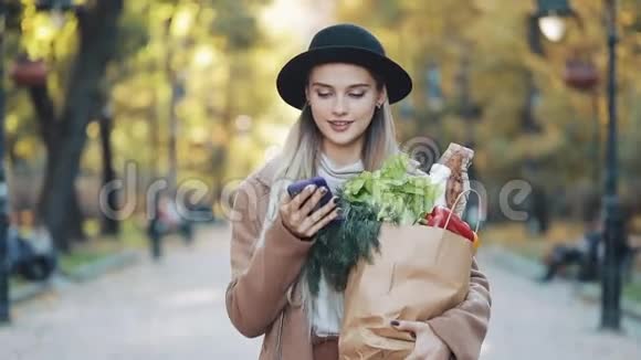 年轻漂亮的女人穿着时髦的外套走在街上拿着一包产品用智能手机视频的预览图