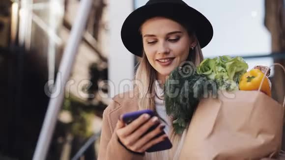 年轻漂亮的女人穿着时髦的外套走在街上拿着一包产品用智能手机视频的预览图