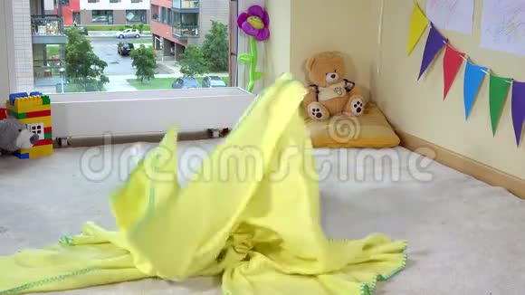淘气的幼儿女孩躲在地板上的黄色格子毯子下面视频的预览图