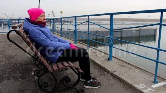 一个女孩坐在码头上的长凳上在寒冷的阴天看着远处视频的预览图