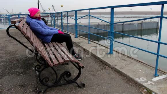 一个女孩坐在码头上的长凳上在寒冷的阴天看着远处视频的预览图