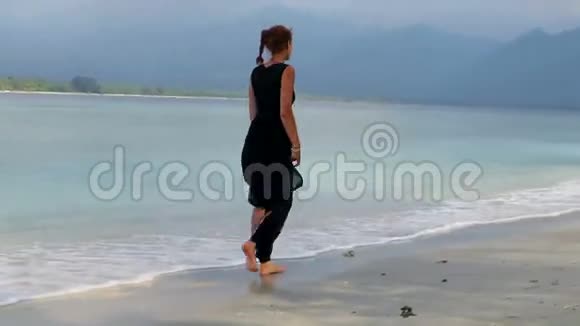 穿着优雅的黑色连衣裙的女人走在美丽的海滩上视频的预览图
