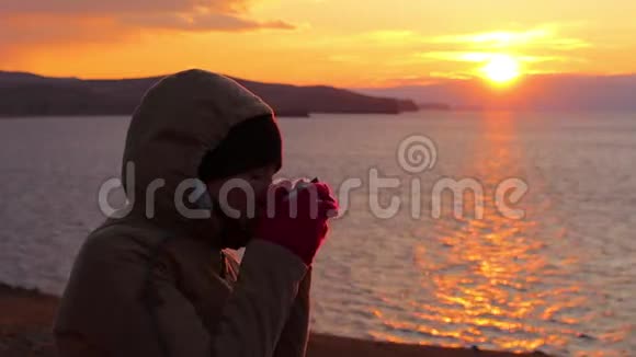 女孩坐在海边喝热茶美丽的金色夕阳视频的预览图