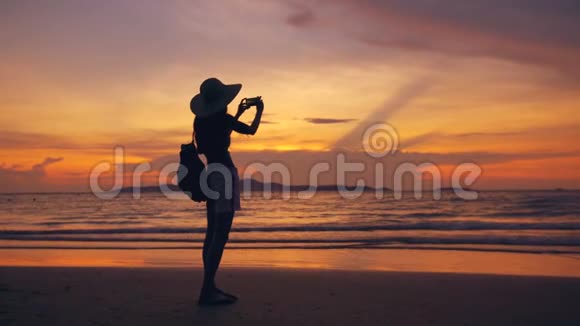 海洋海滩日落时分戴帽子的年轻游客女士用手机拍照剪影视频的预览图