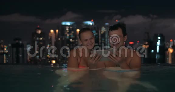美丽的年轻夫妇在游泳池的夜景在摩天大楼屋顶和城市景观的平板上观看视频的预览图