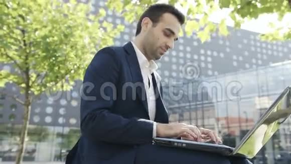 年轻商人坐在办公室附近的公园里用笔记本电脑视频的预览图