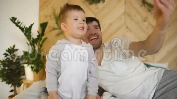 年轻微笑的父亲和儿子在床上用智能手机拍照视频的预览图