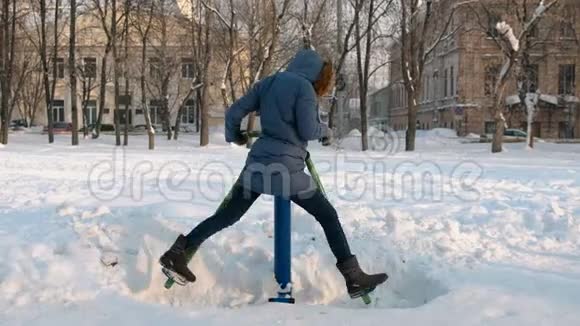 穿着蓝羽绒服带头罩的女人正在城市冬季公园的模拟器上做腿部运动视频的预览图