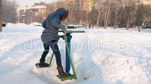 穿着蓝羽绒服的女人带着头罩在城市的一个冬季公园里用模拟器做腿部运动视频的预览图