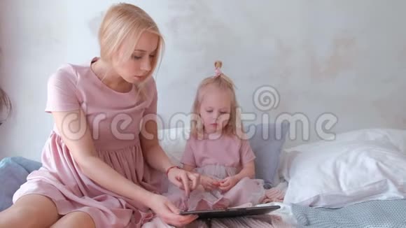妈妈和女儿看平板电脑年轻迷人的金发女郎和她迷人的小女儿穿着粉红色的裙子看着视频的预览图