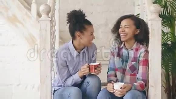两个非洲裔美国卷毛女孩坐在楼梯上在家里一起欢笑和聊天视频的预览图