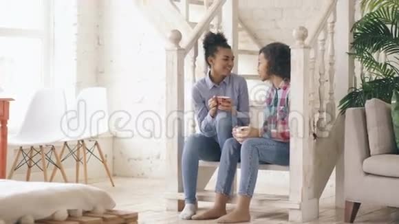 两个非洲裔美国卷毛女孩坐在楼梯上在家里一起欢笑和聊天视频的预览图
