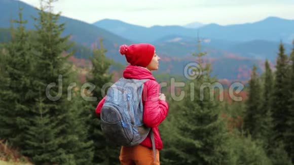 女徒步旅行者带着背包穿着红色夹克和橙色裤子站在山上和森林的背景上视频的预览图