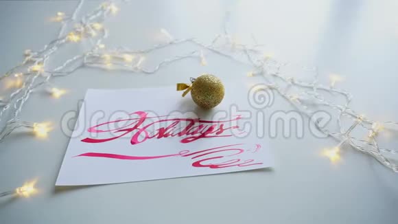 带有红色文字节日的刻字卡放在桌子上的白纸上上面有闪烁的花环和圣诞球视频的预览图