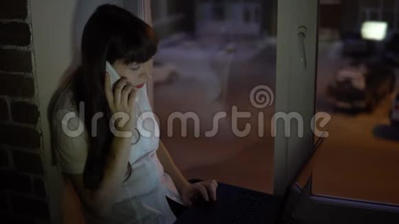 一个女商人坐在靠近一扇黑暗窗户的窗台上在笔记本电脑上工作视频的预览图