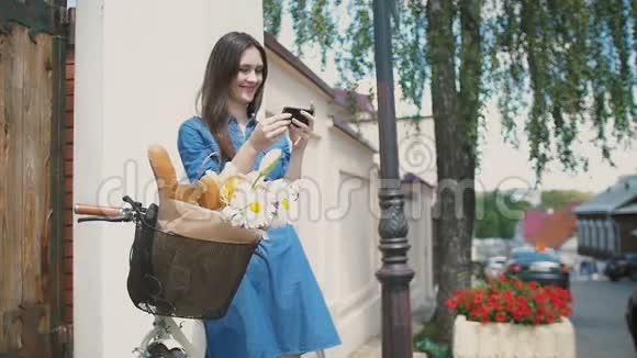 布鲁内特带着微笑的女孩用她的智能手机站在大楼附近骑着一辆自行车篮子里放着花慢悠悠地走着视频的预览图