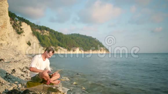 快乐的男人带着现代笔记本电脑坐在海滩上远程工作视频的预览图
