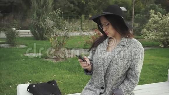 戴着黑帽子戴着眼镜的漂亮年轻女人在城市公园用智能手机视频的预览图