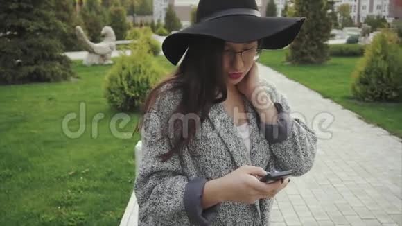 戴着黑帽子戴着眼镜的漂亮年轻女人在城市公园用智能手机视频的预览图