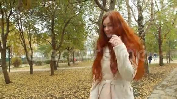 穿着白色大衣的女孩留着红色的长发扫着秋公园的林荫道视频的预览图