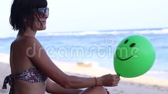 年轻的女人兴高采烈地坐在沙滩上拿着绿色的气球印度尼西亚巴厘全高清50fps在海洋附近视频的预览图