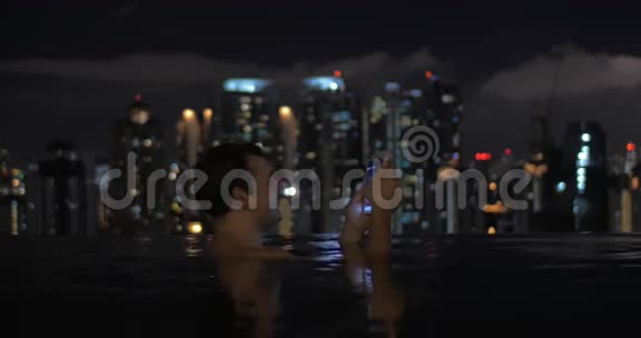 在摩天大楼屋顶上的游泳池中的人的景观使用平板电脑对抗夜间城市景观马来西亚吉隆坡视频的预览图