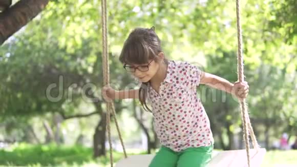 小女孩在夏天公园的秋千上荡秋千在附着在树上的绳子上摆动快乐无忧无虑的童年慢慢慢慢视频的预览图