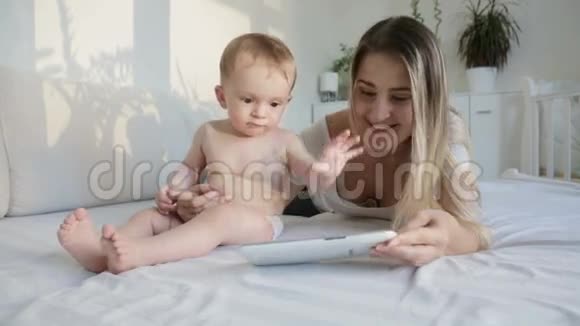 美丽的年轻母亲和她的小儿子在卧室里看数码平板上的卡通片视频的预览图