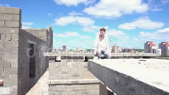 穿着白色运动衫的年轻红发男子坐在屋顶的砖墙上视频的预览图